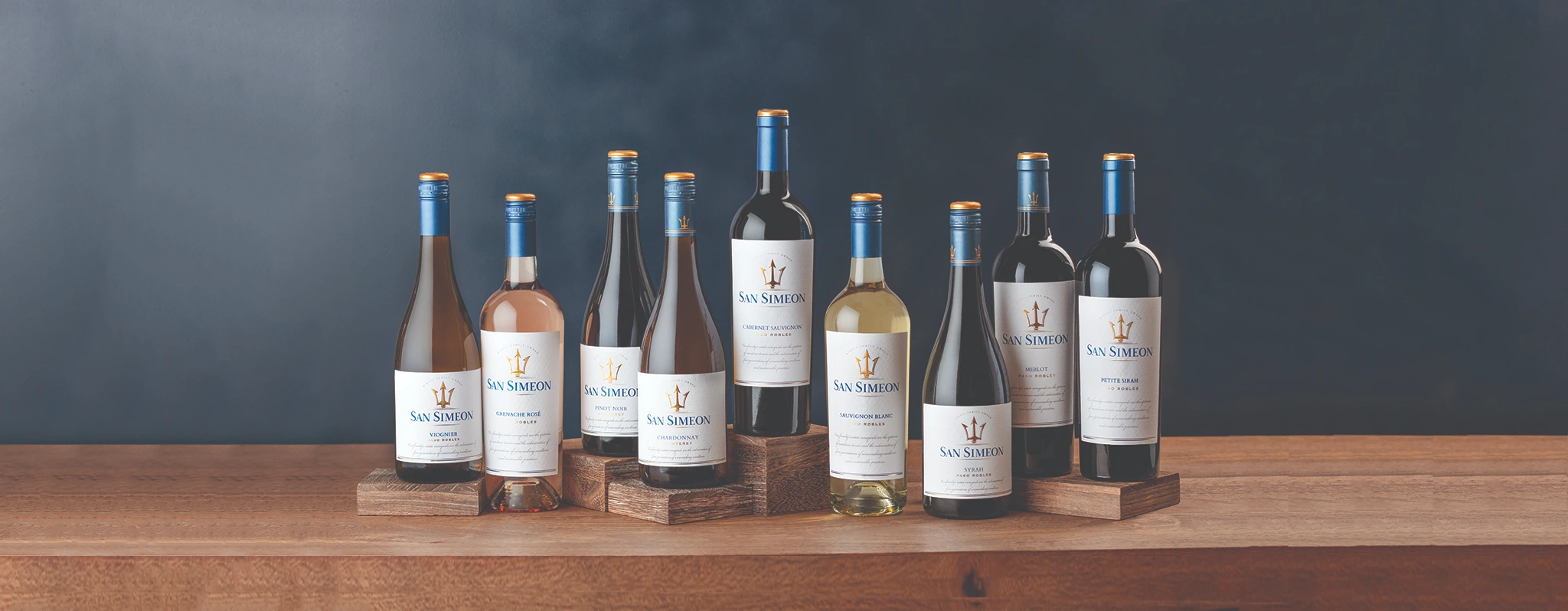 A variety of San Simeon wine bottles displayed on a wooden table against a blue background.